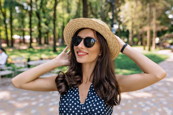 Закрыть наружный портрет милой дружелюбной стильной женщины в солнцезащитных очках и шляпе в летнем платье держит шляпу Стоковое Фото