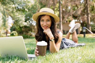 Siyah saçlı, sevimli bir kız. Şapka ve elbise giyiyor. Çimlerin üzerinde mutlu bir gülümsemeyle dizüstü bilgisayar ve kahveyle yatıyor.