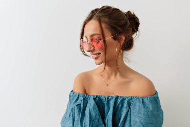 Güzel gülüşü olan sevimli beyaz kadın beyaz arka plana tepeden bakıyor. Koyu renk saçlı kız yazlık kot pantolon ve pembe gözlük takıyor. Duygusallık ve kadınlık kavramı