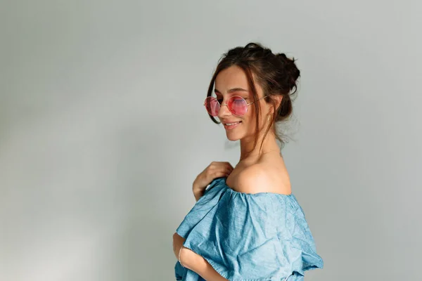 Beautiful lady in pink glasses and denim top is looking down and smiling on isolated background. Brown-haired enjoying happy time at home. Emotion concept, facial expression.