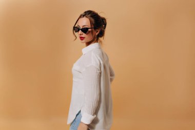 Kafasında kiriş olan şık, sevimli bayan beyaz gömlek giyiyor koyu renk gözlüklü, kırmızı rujlu, arka planda bej duvarda kameraya poz veriyor. Moda kadını fotoğraf çekiminde poz veriyor.