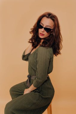 Zarif şık takım elbiseli, gözlüklü, dalgalı siyah saçlı şık bir kadın bej arka planda sandalyede oturuyor. Güzel kız saçına dokunuyor ve fotoğraf çekiminde poz veriyor.