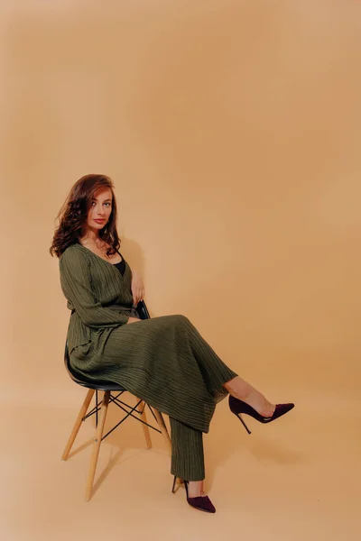 Elegant incredible woman with dark hair is dressed dark green suit and heels and sitting on chair over isolated background. Pretty tanned lady in wide pants and jacket is looking at camera