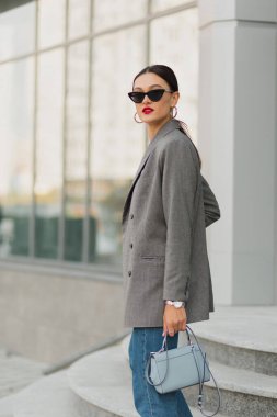 Siyah saçlı, gri ceketli, kot pantolonlu ve güneş gözlüklü havalı Avrupalı kız dışarıda kameraya bakıyor. Gözlüklü şık kadın dışarıda oturuyor.