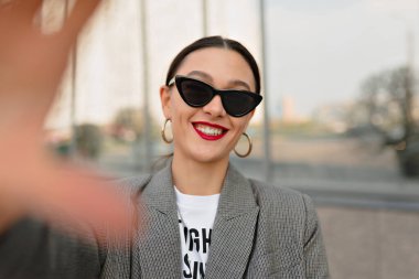 Çekici gülüşü olan güzel, şık esmer kız siyah güneş gözlüğü takıyor ve kırmızı dudakları dışarıda selfie çekiyor. Mutlu gülümseyen kadın şehirde dinleniyor.