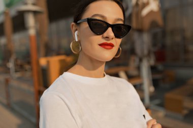 Koyu saçlı, kırmızı dudaklı, koyu renk gözlüklü ve beyaz tişörtlü inanılmaz çekici bir bayan güneş ışığında kameraya gülümsüyor. Şehirde güzel bir kızın dış portresi.