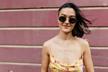 Mutlu güzel model dışarıda pembe duvarın üzerinde poz verirken kameraya bakıyor. Saçları rüzgarda uçuşuyor, yaz elbisesi giyiyor. Açık hava fotoğrafı.