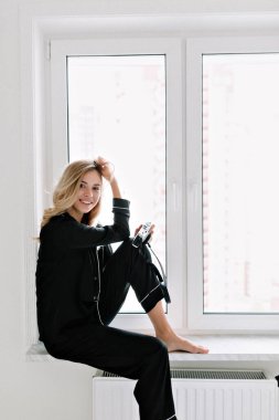 Siyah takım elbiseli şık güzel bir kızın tam boy portresi pencerede oturuyor ve kameraya gülümsüyor. Güneş ışığında odasında dinlenen güzel kız. 