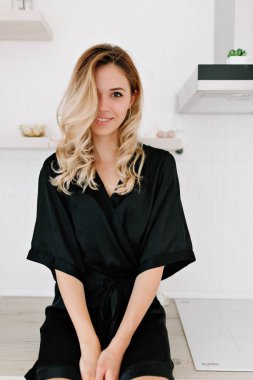 Dalgalı sarı saçlı güzel kız mutfakta masada oturuyor ve kameraya gülümsüyor. Siyah elbiseli, yüzü saçla kaplı sevimli bir kadın.