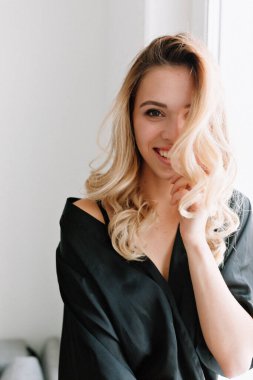 Düzgün gülümseyen bir kadın koyu renk bluz giyer ve pencerenin yanında zevkle poz verir. Dairesinde bir fincan kahveyle oturan kıvırcık kadın manken.