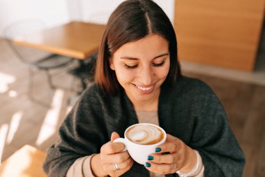 Güzel gülüşlü zarif bayan elinde kahve tutuyor ve dinleniyor. Bronzlaşmış tenli ve koyu renkli manikürlü Avrupalı bir kadın güneşli bir sabahta kahvaltı yapıyor. 