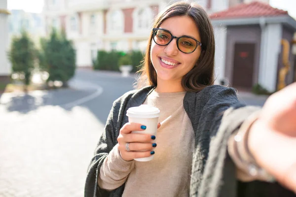 Gözlüklü, siyah saçlı mutlu zarif kadın elinde kahve tutuyor ve şehrin güneşli caddesinde selfie çekiyor. Yüksek kalite fotoğraf