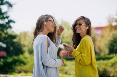 Parlak elbiseli, uzun dalgalı saçlı güzel modern kadınlar şehrin arka planında güneşli ve sıcak bir günde müzik dinliyorlar. İki muhteşem kız kardeşin açık hava portresi.