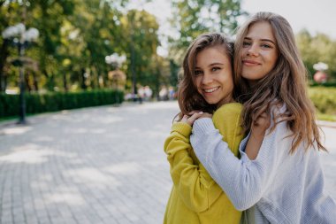 Uzun dalgalı saçlı, parlak kazak giyen iki kız gülümsüyor ve dışarıda vakit geçiriyor. Beyaz elbiseli, güz günü arkadaşını kucaklayan neşeli, beyaz bir kadın..