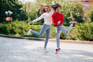 Parlak kazaklı ve kotlu iki enerjik kızın tam boy portresi yeşil güneşli parkın arka planında zıplıyor ve gülüyor. Yüksek kalite fotoğraf