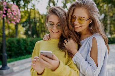 Uzun saçlı, yuvarlak gözlüklü ve sarı kazaklı güzel bir bayanın portresi dışarıda kız kardeşiyle birlikte kulaklıkla müzik dinliyor. İki kız akıllı telefonları kaydırıyor. 