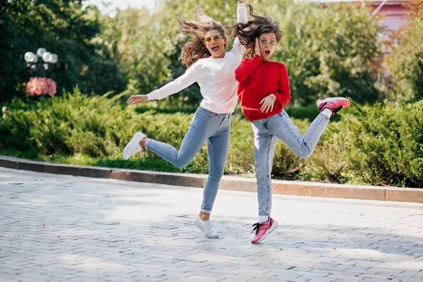 Parlak kazaklı ve kotlu iki enerjik kızın tam boy portresi yeşil güneşli parkın arka planında zıplıyor ve gülüyor. Yüksek kalite fotoğraf