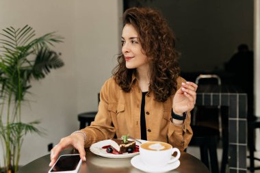 Yumuşak bluz giymiş kıvırcık saçlı bir kadın modern kafenin arka planında bir fincan kahve ve tatlı ile oturuyor..