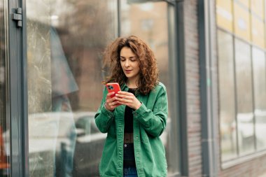Yeşil ceketli, şık, modern Avrupalı kadın akıllı telefonuyla sokakta yürüyor. Güzel bayan dışarıda vakit geçiriyor.