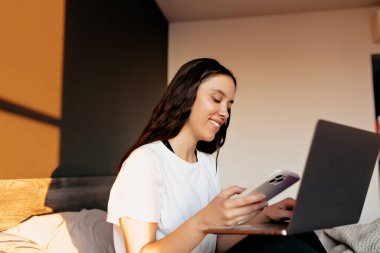 Smiling attractive pretty woman with happy smile in white shirt is sitting on the bed in sunlight and typing on laptop. Girl is holding zoom conference on laptop with phone. High quality photo