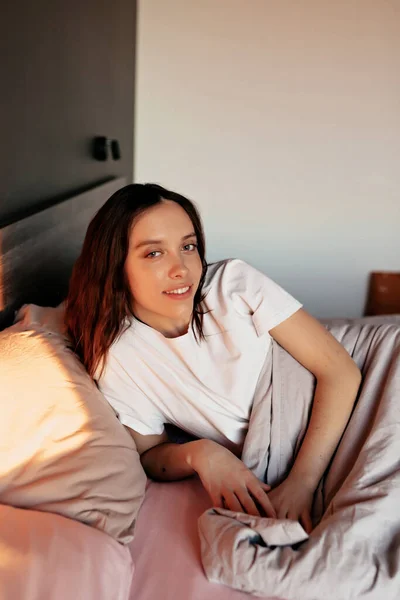 可爱可爱的姑娘 一头乌黑的波浪般的头发 早上醒来 躺在床上 带着幸福的笑容 高质量的照片 早上在现代公寓床上躺着的漂亮女孩的画像 — 图库照片
