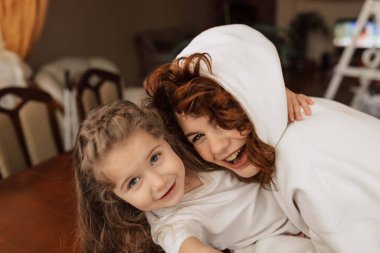 Close-up portrait of young mother with curls wearing hoodie is hugging her little daughter. Woman with smile looks at child. High quality photo