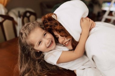 Uzun dalgalı saçlı sevimli küçük bir kız annesine sarılıyor ve evde iyi vakit geçiriyor. Kıvırcık kadın küçük kızını sevgiyle öpüyor. Yüksek kalite fotoğraf