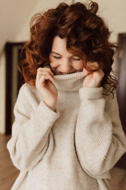 Charming cute lady with curls wearing knitted pullover is laughing and looking down with closed eyes. Indoor portrait of romantic pretty lady. High quality photo
