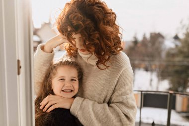 Happy excited little girl with long wavy hair is smiling and spend time at home with her mother. Pretty woman with her little daughter posing in sunlight on balcony. High quality photo