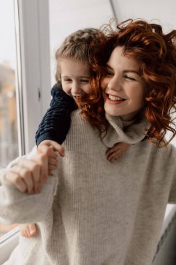 Indoor portrait of young woman with red curly hair dressed knitted pullover is playing with her little daughter and laughing at home. High quality photo