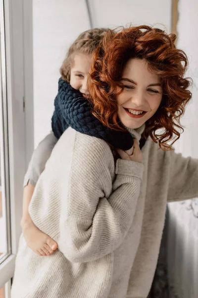 Bukleli örgü kazaklı mutlu hamile kadın küçük kızının elini tutuyor ve güneşli bir günde evde birlikte eğleniyor. Yüksek kalite fotoğraf