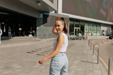 Saçları toplayan çekici Avrupalı kadın arkadan poz veriyor. İnanılmaz beyaz kızın açık hava portresi sıcak güneşli bir günde kot pantolon ve tişört giyer.