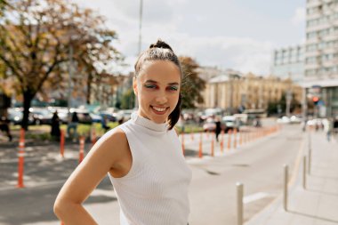 Güneşli bir günde dışarıda gülümseyen zarif, şık bir bayan. Parlak makyajlı, açık renk tişörtlü sevimli genç bir kız şehrin arka planına bakıyor.