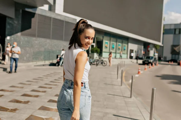Beyaz tişörtlü ve kot pantolonlu harika şık bayan şehir merkezinde güneşli bir günde şehirde dolaşıyor. Yüksek kalite fotoğraf