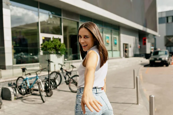 Parlak makyajlı, beyaz tişörtlü ve kot pantolonlu büyüleyici ve mutlu kız kameraya doğru dönüyor ve şehirde yürürken el sallıyor. Yüksek kalite fotoğraf