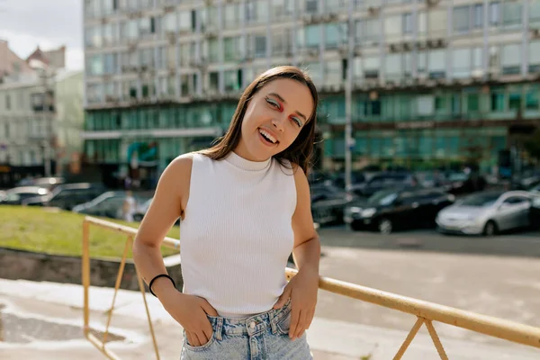 Uzun kahverengi saçlı ve makyajlı çekici genç bayan beyaz tişört giyiyor ve güneşli bir günde şehirde yürürken kotlar kameraya poz veriyor. Yüksek kalite fotoğraf