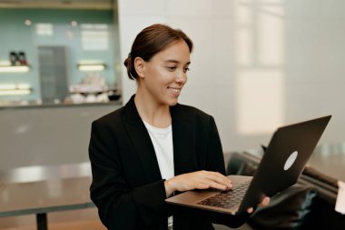 Koyu renk ceketli iş kadını rahat ofisinde güneş ışığında dizüstü bilgisayarda çalışıyor. Ofisteki kadın portresi