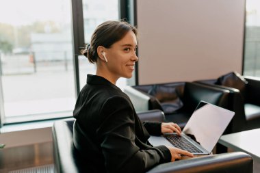 Kapalı alanda, koyu renk ceketli, kulaklıklı sevimli iş kadınının portresi modern şık ışık ofisinde dizüstü bilgisayarda çalışıyor.