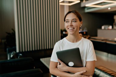 Siyah saçlı çekici genç bayan elinde dizüstü bilgisayarla modern şık ofisin arka planında harika bir gülümsemeyle beyaz gömlekli. 
