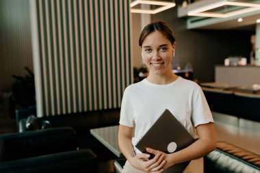 Siyah saçlı, beyaz tişörtlü, bej pantolonlu, çok güzel bir bayan elinde dizüstü bilgisayarla kameraya bakıyor.