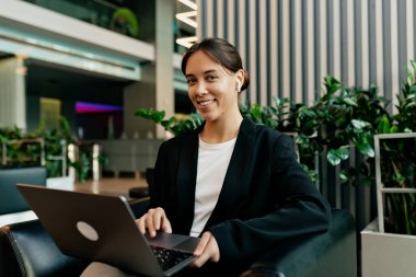 Gündüz kıyafetleri içindeki çekici bayan güneşli bir günde ofiste dizüstü bilgisayarla çalışıyor. Genç bir kadın laptopta çalışıyor ve kablosuz kulaklıkla müzik dinliyor.