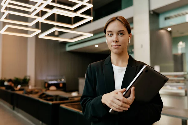 Kendine güveni tam, koyu renk ceketli güzel bir kadın dizüstü bilgisayar tutuyor ve modern şık ofisin arka planında harika bir gülümsemeyle kameraya poz veriyor.