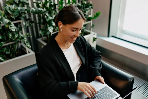 Beyaz tişörtlü, siyah ceketli, kulaklıklı hoş bir bayan modern modaya uygun bir ofiste dizüstü bilgisayarda yazıyor.