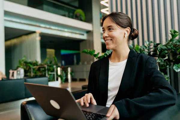 Şık, modern, kulaklıklı, ceketli, gülümseyen, dizüstü bilgisayarda yazan ve ofiste poz veren esmer kadın. Kadın işçi ofiste çalışıyor.