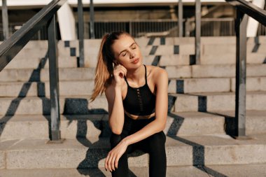 Spor giyimli, çekici genç bir kadın sabahın köründe spor meydanında gündoğumunun tadını çıkarıyor. Neşeli ruh hali, gerçek duygular, sağlıklı yaşam tarzı, kapalı gözlerle gülümseme.