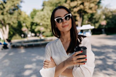 Elegant stylish woman with straight dark hair wearing sunglasses and white shirt is holding cup with coffee and posing at camera in sunlight in green summer park in the city. High quality photo