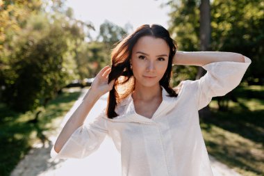 Bluz dışarıda güneş ışığında poz verirken güzel genç bir kadının portresini yakından çek. Kahverengi gözlü esmer, bol saçlı yeşil güneşli parkta yürüyor. 