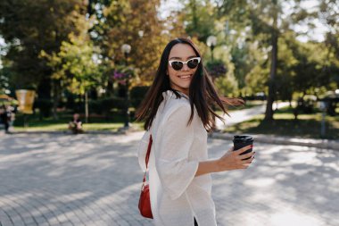 Excited girl with wonderful smile wearing white shirt in sunglasses is holding coffee is turn around at camera while walking in sunny green park in the city. High quality photo
