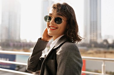 Gülümseyen, kırmızı dudaklı, güneş gözlüklü, gri ceketli mutlu esmer kadının profil portresi ılık bir bahar gününde şehir dışında yürüyor. Yüksek kalite fotoğraf