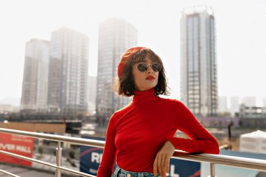 Güneş gözlüklü, kısa koyu renk saçlı, kırmızı gömlekli muhteşem büyüleyici kadın şehrin arka planında güneş ışığında poz veriyor. Yüksek kalite fotoğraf